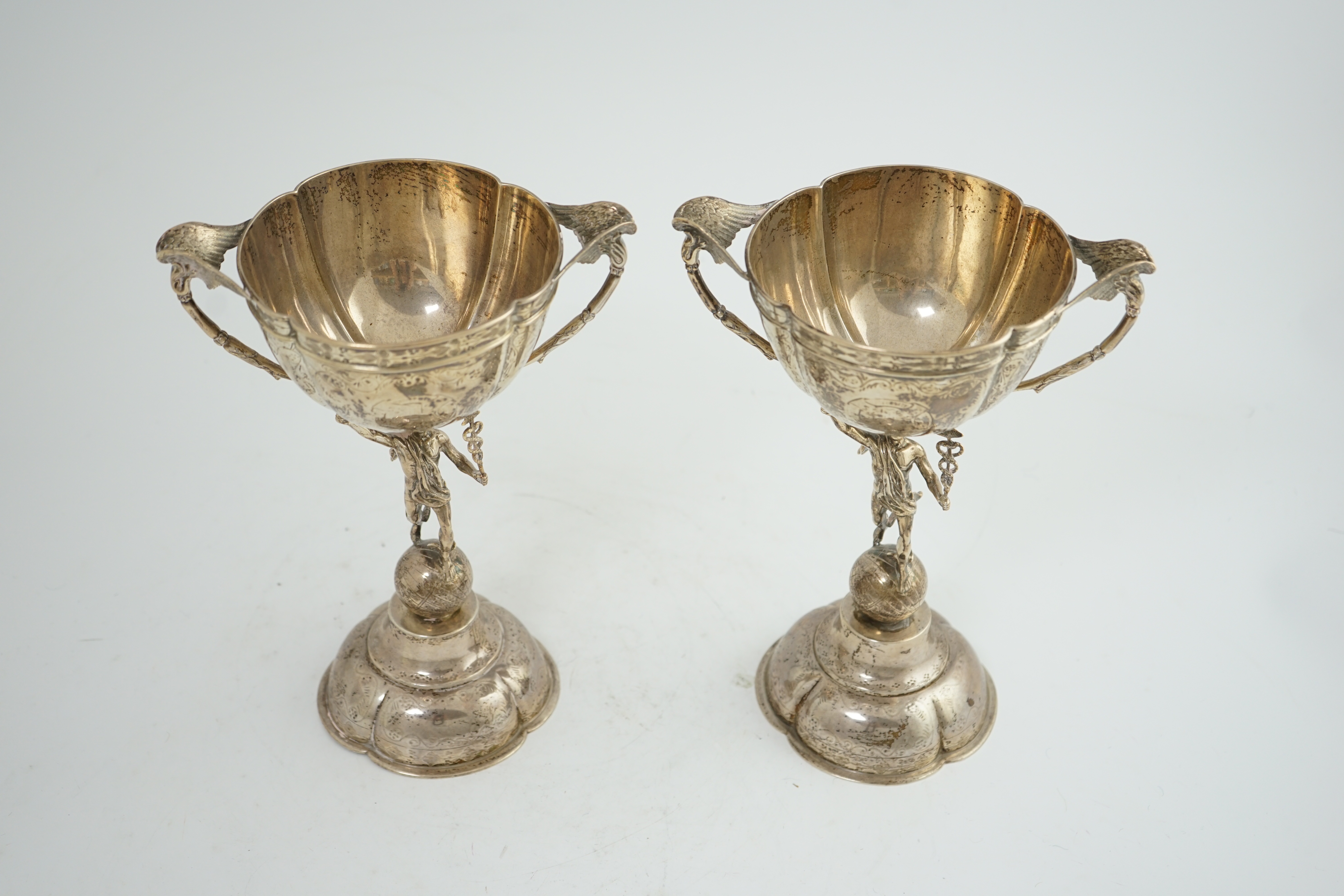 A pair of Victorian silver two handled pedestal cups, each figural stem modelled as Hermes upon a globe, by Edward Charles Brown